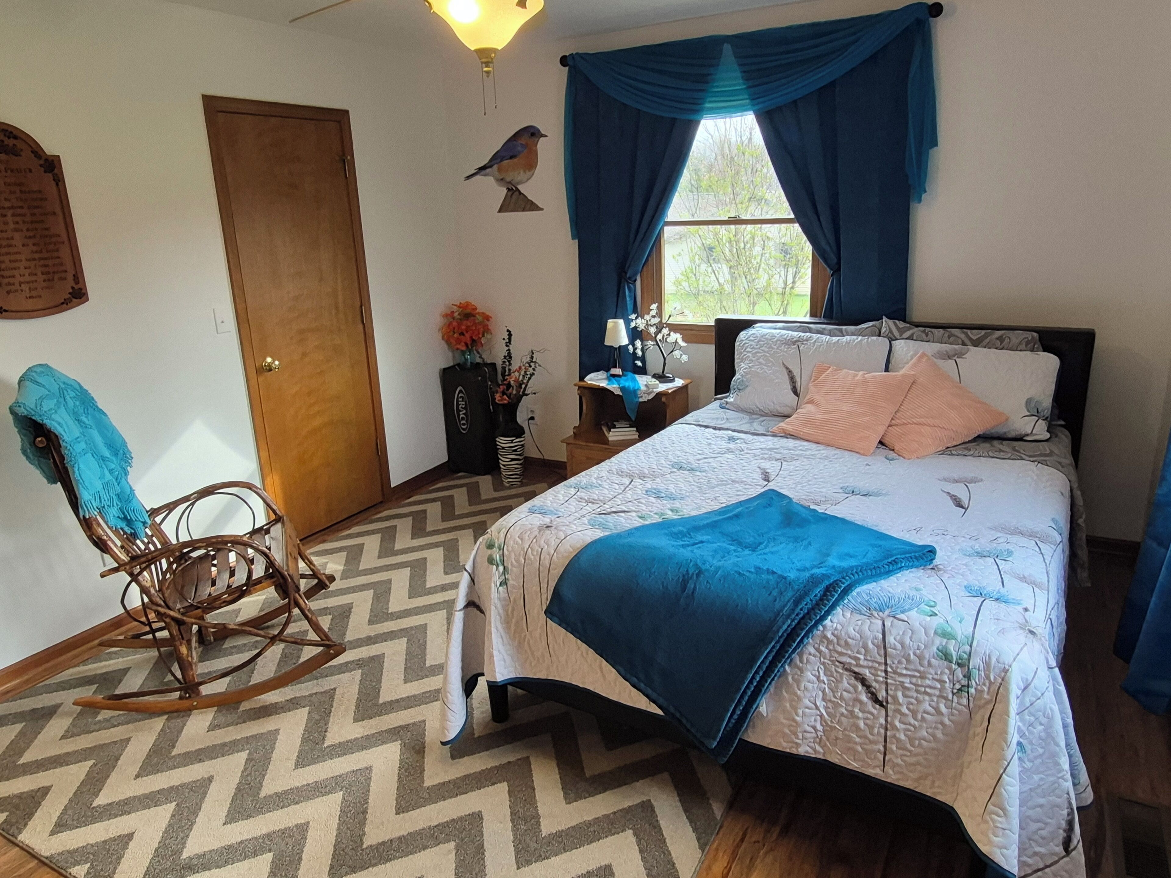 Un dormitorio con cama, una mecedora, un ventanal con cortinas, una escultura de pájaro en la pared y una puerta.