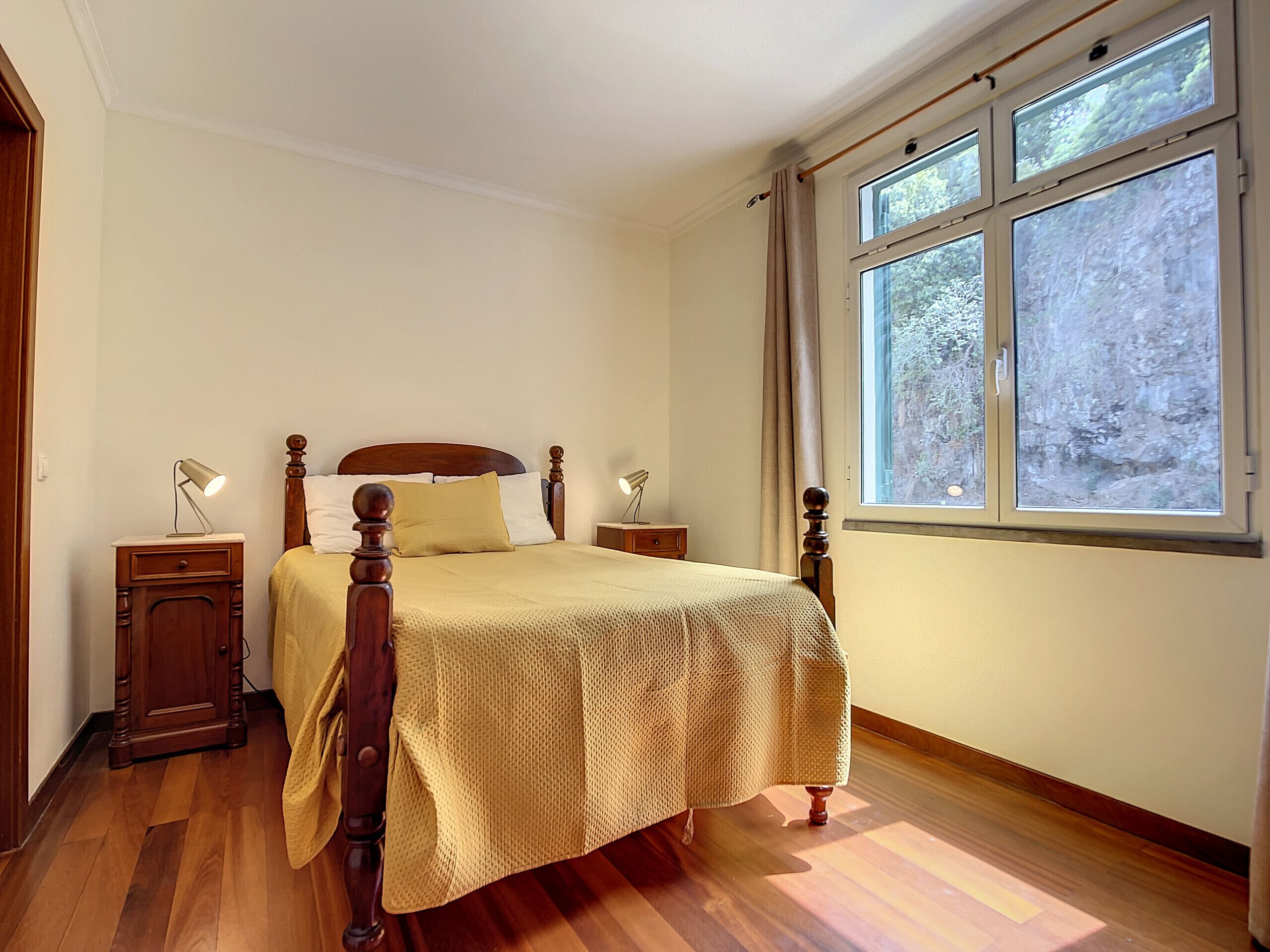 Une chambre à coucher avec un lit en bois, deux tables de chevet, une fenêtre donnant sur des arbres et un plancher en bois.