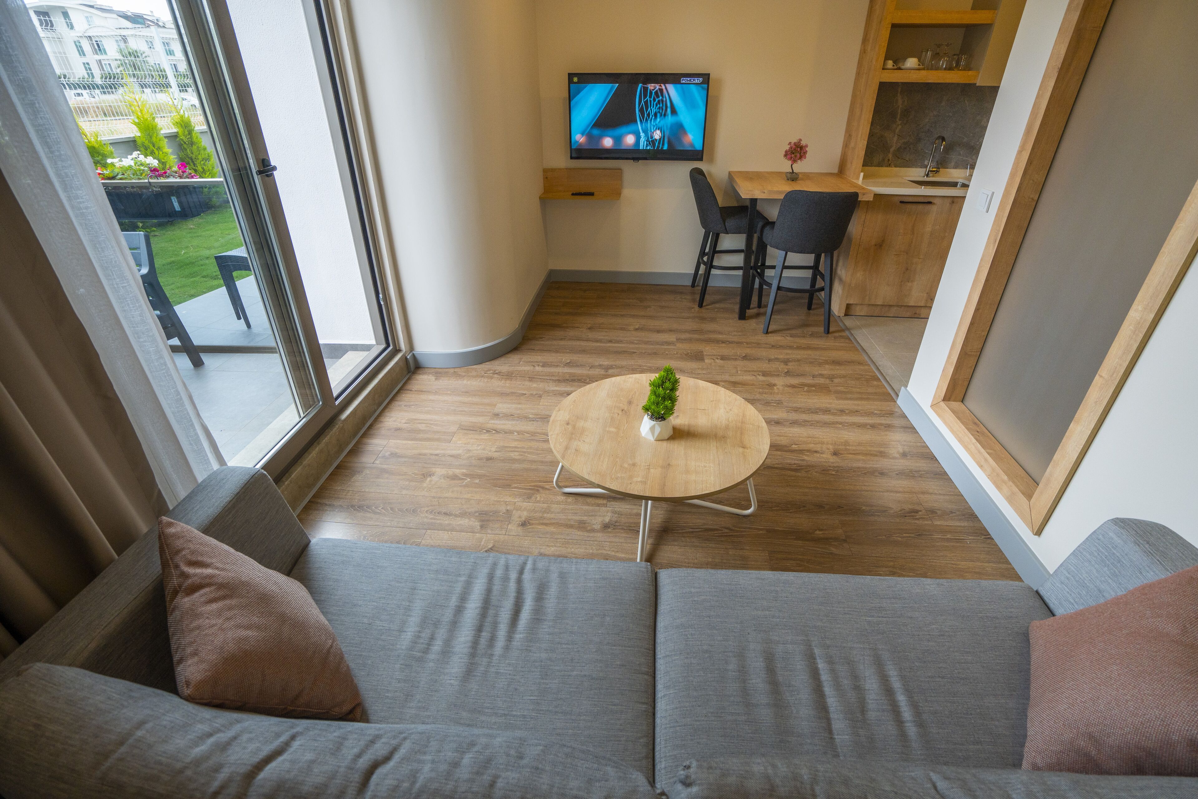 Una sala de estar moderna con un sofá, una mesa redonda con una planta, un televisor montado en la pared y una cocina al fondo.