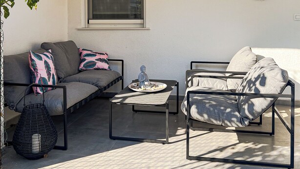 A sunlit patio with a sofa, armchair, and table, featuring a window, decorative wall pieces, and a hanging plant.
