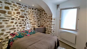 Ein Schlafzimmer mit Steinwand, einem Bett mit floralem Überwurf, einem Nachttisch, einem kleinen Tisch mit Lampe und einem Fenster mit Aussicht.