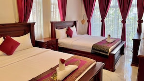 A hotel room with two beds, each with a wooden headboard, white bedding, and red pillows. The room has large windows with red curtains, a wooden nightstand, and a small table with a lamp.