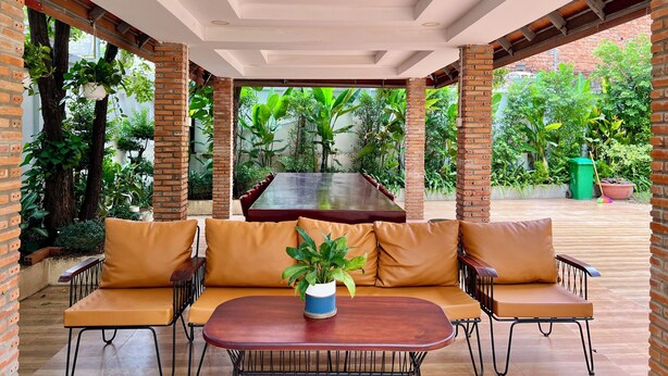 A covered outdoor seating area with a wooden table and cushioned chairs, surrounded by brick walls and lush greenery.