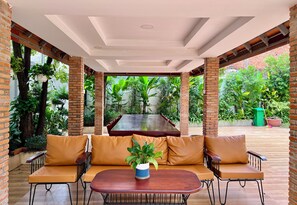 A covered outdoor seating area with a wooden table and cushioned chairs, surrounded by brick walls and lush greenery. - NyNa Villa Kampot (Kampot)