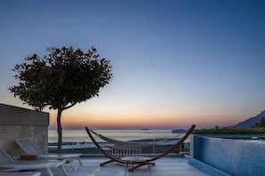 Uma cadeira de rede e espreguiçadeiras em um terraço com vista para uma piscina e um pôr do sol.