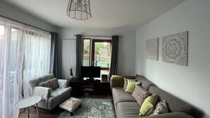 Een woonkamer met een bank, een salontafel en een televisie. Er hangen gordijnen voor de ramen, en een decoratief kunstwerk is zichtbaar.