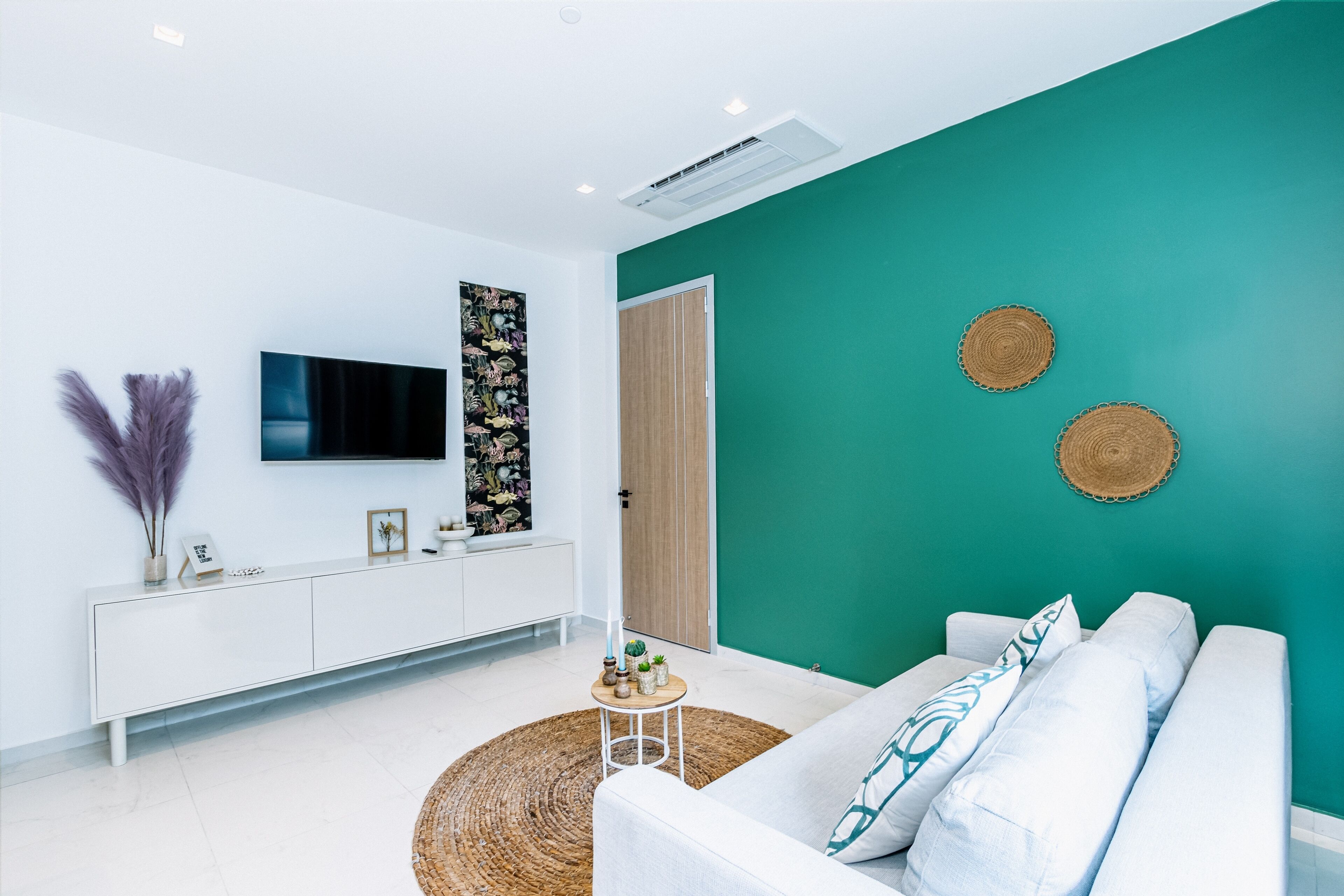 A modern living room with a teal accent wall, white furniture, a flat-screen TV, and decorative items.