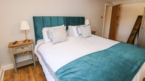 A neatly made bed with a teal headboard, white bedding, and patterned pillows. There is a bedside table with a lamp and a remote control. - The Coach House At The Gables (Shrewsbury)