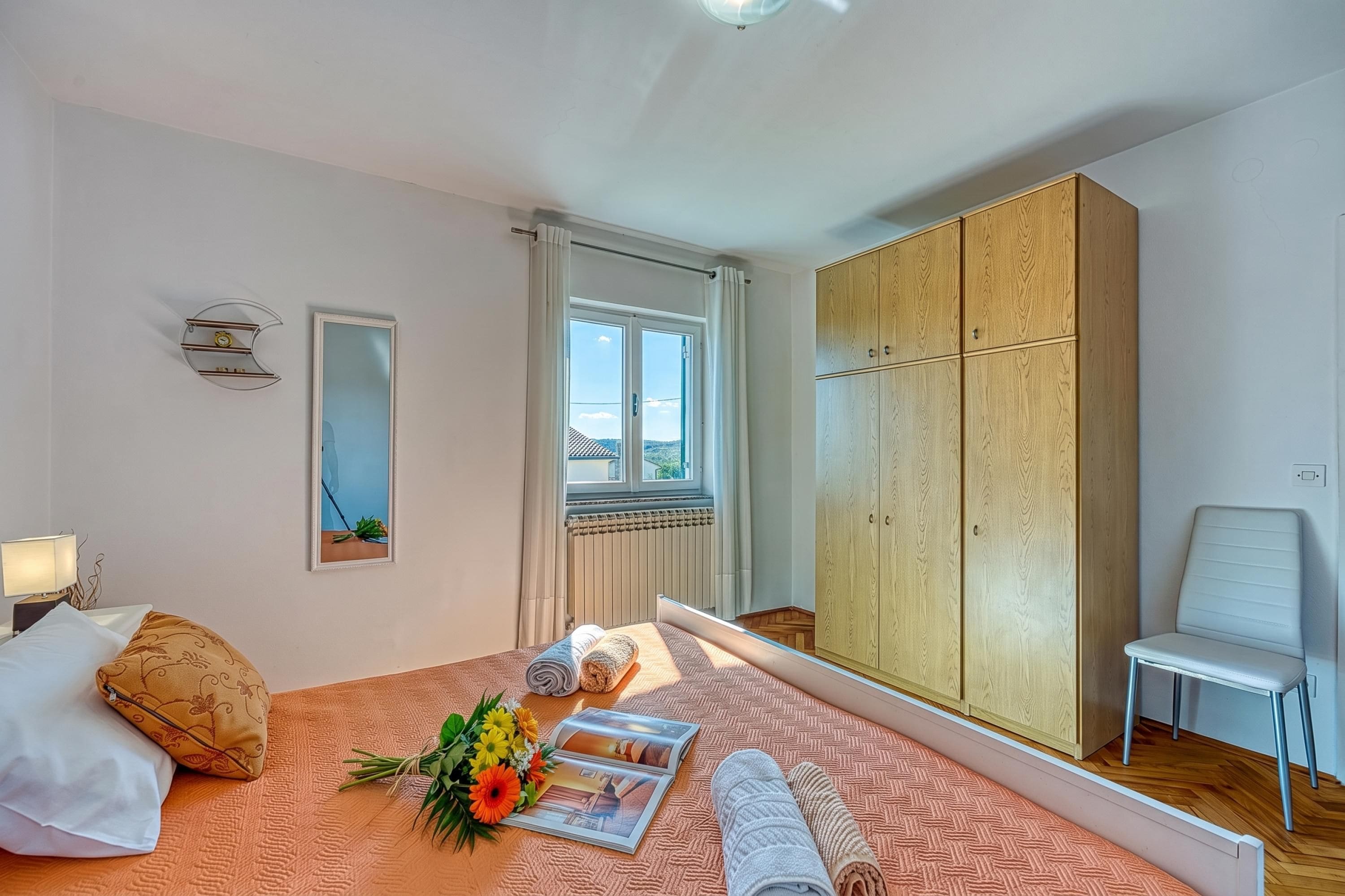 A hotel room with a bed, a bedside table with a lamp, a large wardrobe, a window, and a chair.