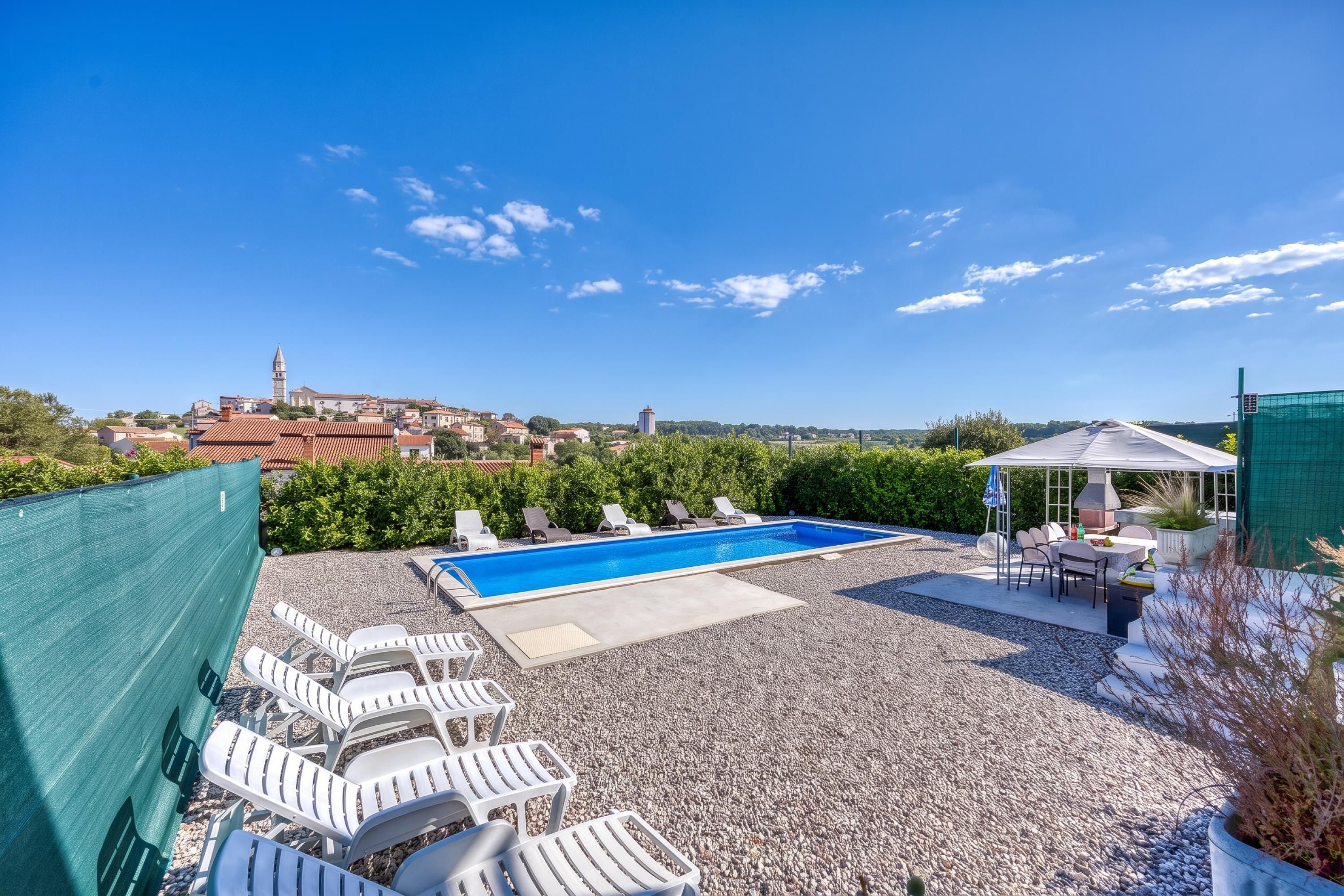 Una piscina al terrat amb gandules, zona de barbacoa i vista a una vila amb una torre de l'església.