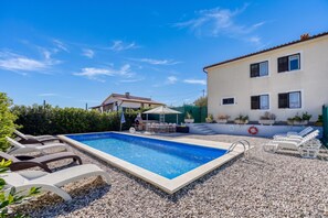 Una piscina con sillones de descanso y zona de barbacoa, rodeada de suelo cubierto de guijarros.
