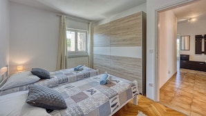 Un dormitorio con dos camas, un ventanal con cortinas, un panel de pared de madera y un baño visible a través de una puerta abierta.