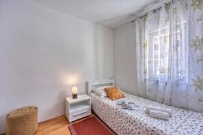 A bedroom with a single bed, a bedside table with a lamp, a window with floral curtains, and a small wooden stool. - Pool House Lilly (Visnjan)