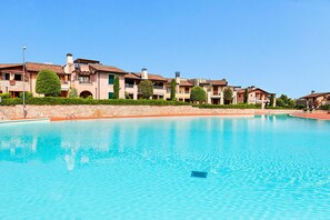 A swimming pool with a clear blue water surface, surrounded by multi-story buildings with balconies and a well-maintained hedge. - La Rocca 2 11 by Wonderful Italy (Manerba del Garda)