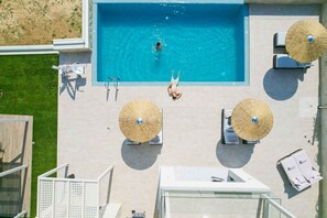 Vue aérienne d’un espace piscine avec des chaises longues et des parasols, entouré d’une pelouse bien entretenue et d’un bâtiment.
