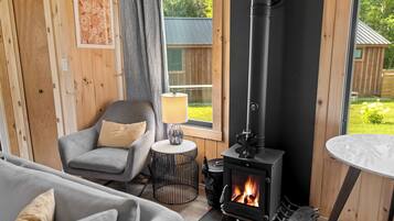 A cozy room with a wood-burning stove, a grey sofa, a small round table with a lamp, and a window overlooking a garden.