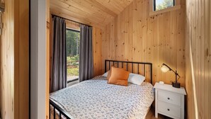 Ein Schlafzimmer mit Holzwänden, einem Bett, Vorhängen, einem Nachttisch mit Lampe und einem Fenster.