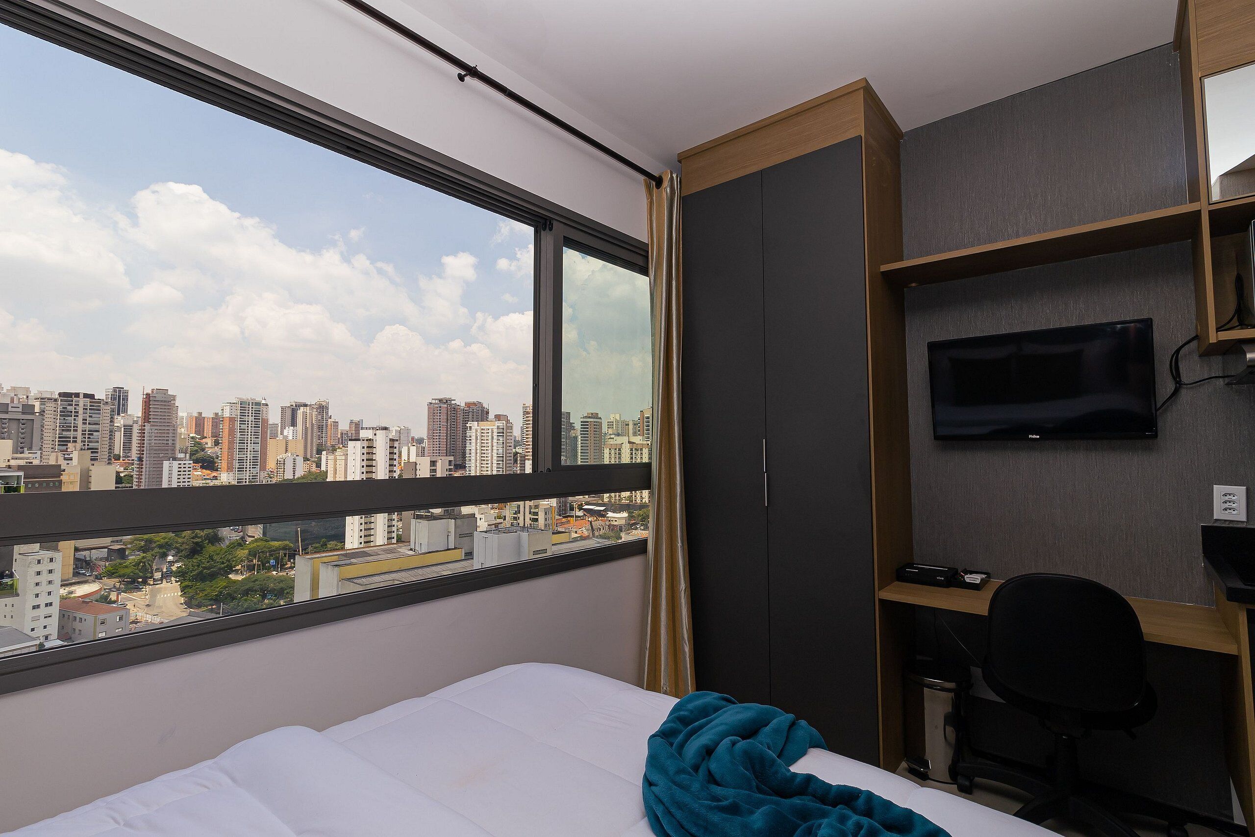 Ein Hotelzimmer mit großem Fenster, einem Bett, einem Schreibtisch mit Stuhl, einem Fernseher und einem Kleiderschrank.
