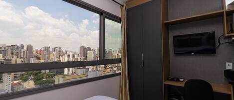 A hotel room with a large window, a bed, a desk with a chair, a TV, and a wardrobe.