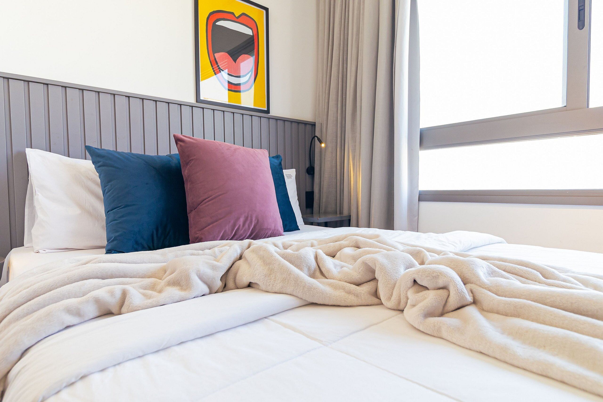 Ein ordentlich bezogenes Bett mit einer beigen Decke, einem weißen Kopfkissen und zwei Kissen in Blau und Rosa.