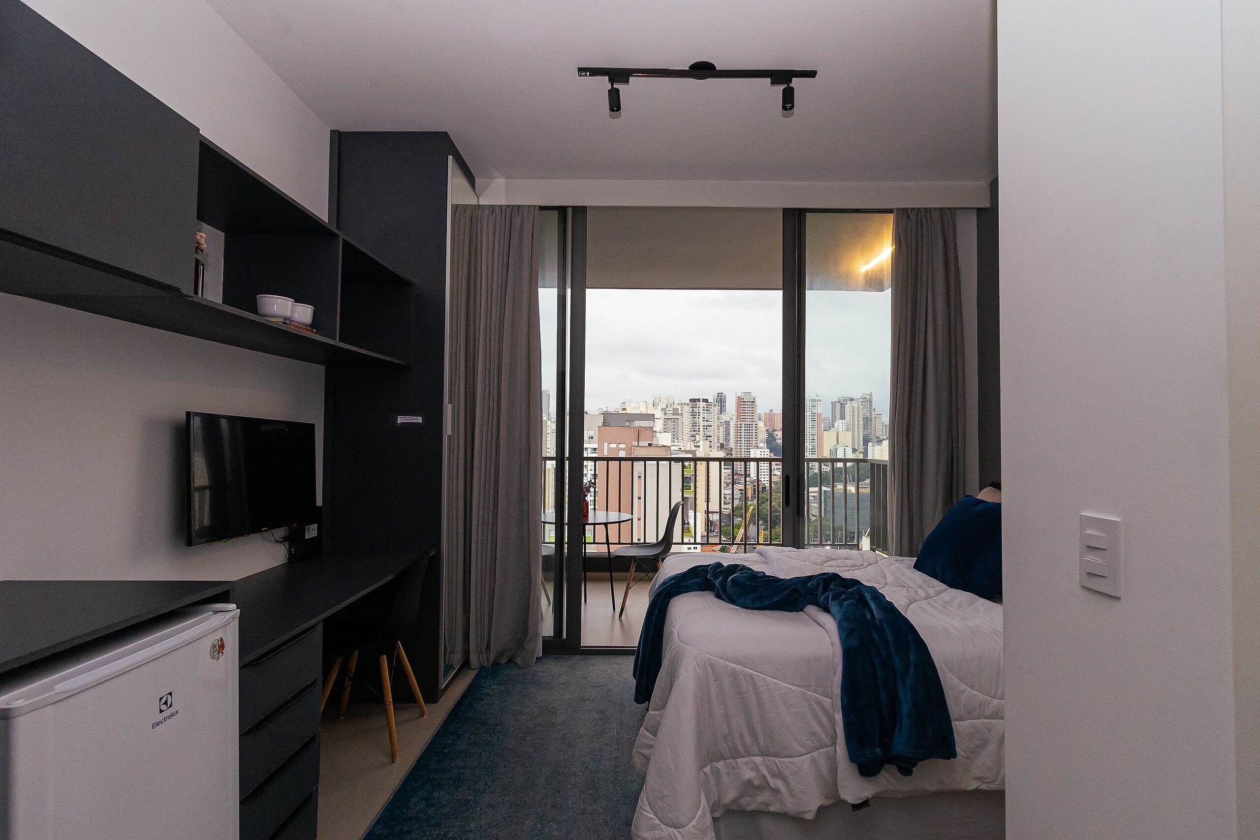 A modern hotel room with a bed, a desk, a television, and a balcony with a city view.