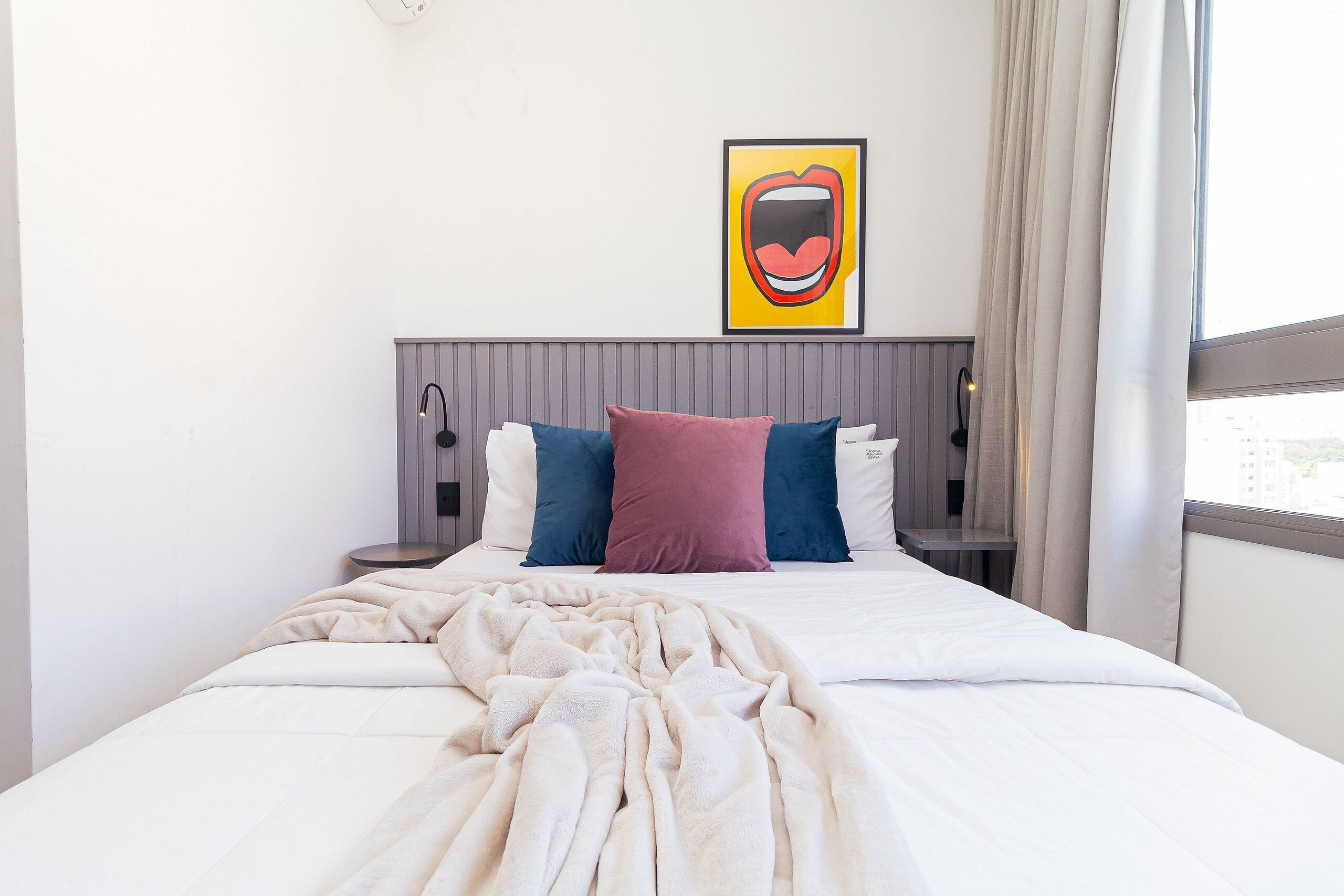 Un lit bien fait, avec une couette blanche et un plaid beige, une tête de lit grise et un ensemble d’oreillers aux couleurs vives.