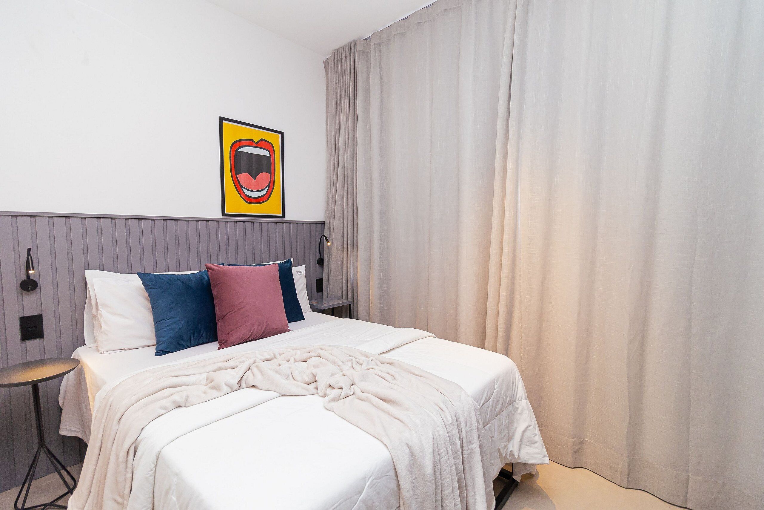Una habitación de hotel con una cama, una mesita de noche, una lámpara de pared y un amplio ventanal con cortinas.