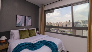 Una habitación de hotel con cama, mesita de noche, lámpara y vistas de la ciudad a través de grandes ventanales.