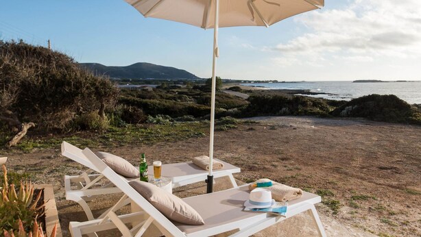 A beachside lounge with a white umbrella, lounge chairs, and a table with a drink and a hat. - Falasarna Seafront House I 50 m to the Beach (Kissamos)