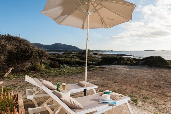 A beachside lounge with a white umbrella, lounge chairs, and a table with a drink and a hat. - Falasarna Seafront House I 50 m to the Beach (Kissamos)
