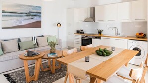 A modern kitchen with white cabinets, a wooden dining table, and a large painting of a beach scene. - Falasarna Seafront House I 50 m to the Beach (Kissamos)