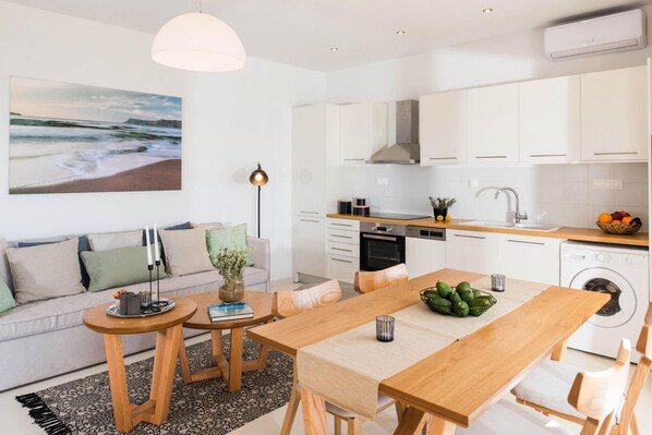 A modern kitchen with white cabinets, a wooden dining table, and a large painting of a beach scene. - Falasarna Seafront House I 50 m to the Beach (Kissamos)
