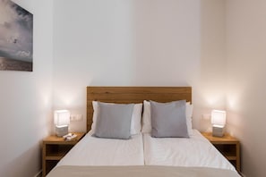 Cama de casal com cabeceira de madeira, duas mesas de cabeceira com abajures e uma tomada na parede.