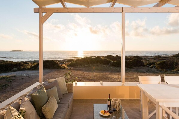 Uma área de estar coberta ao ar livre com vista para o mar, uma mesa com uma garrafa e copos, e um sofá com almofadas.