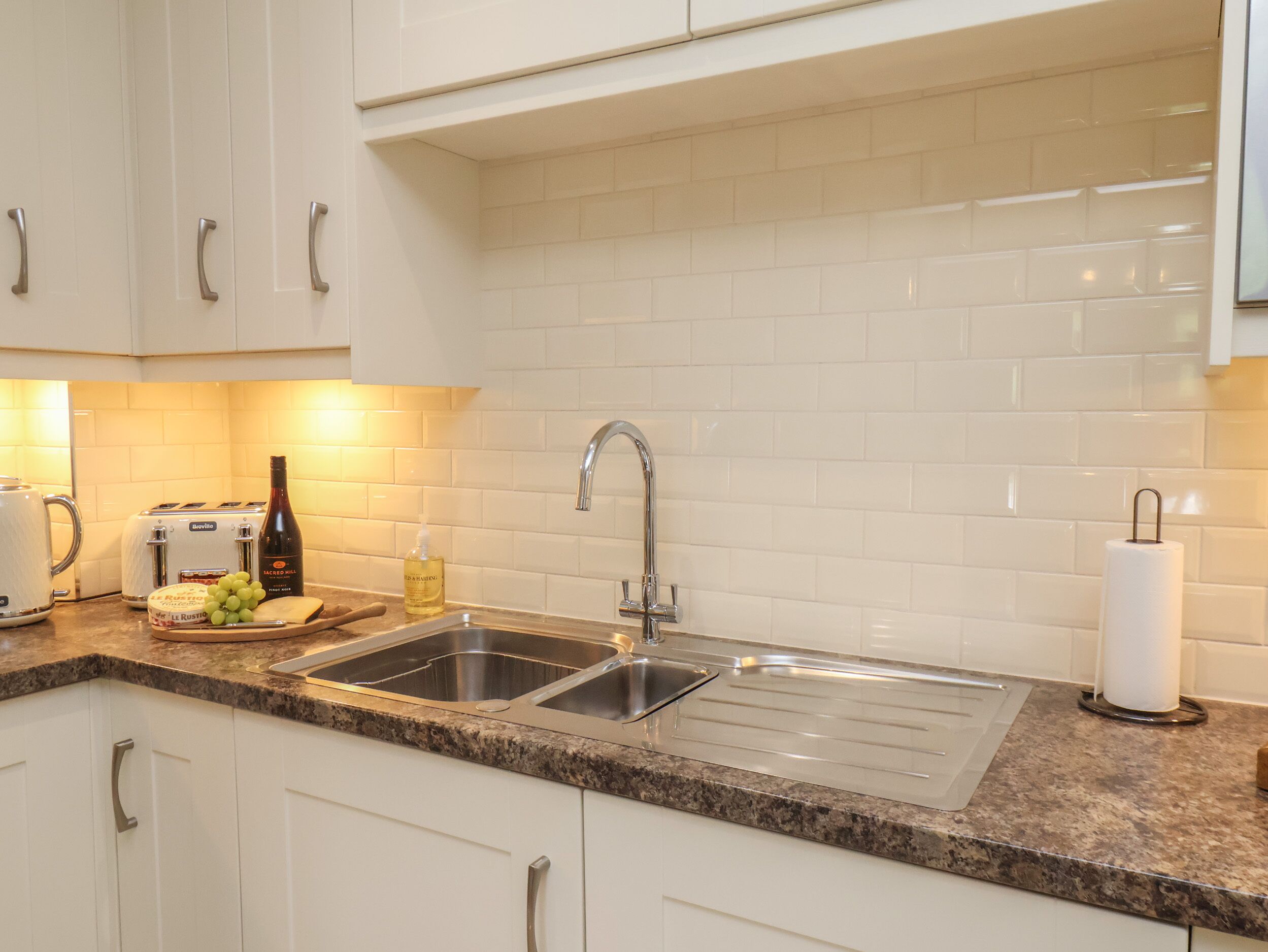 A modern kitchen with white cabinets, a granite countertop, a double sink, a toaster, a bottle of olive oil, and a roll of paper towels.