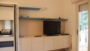A room with a TV on a stand, a shelf with decorative items, and a door leading to an outdoor area. - Lovely Apartment in Lignano Sabbiadoro (Lignano Sabbiadoro)