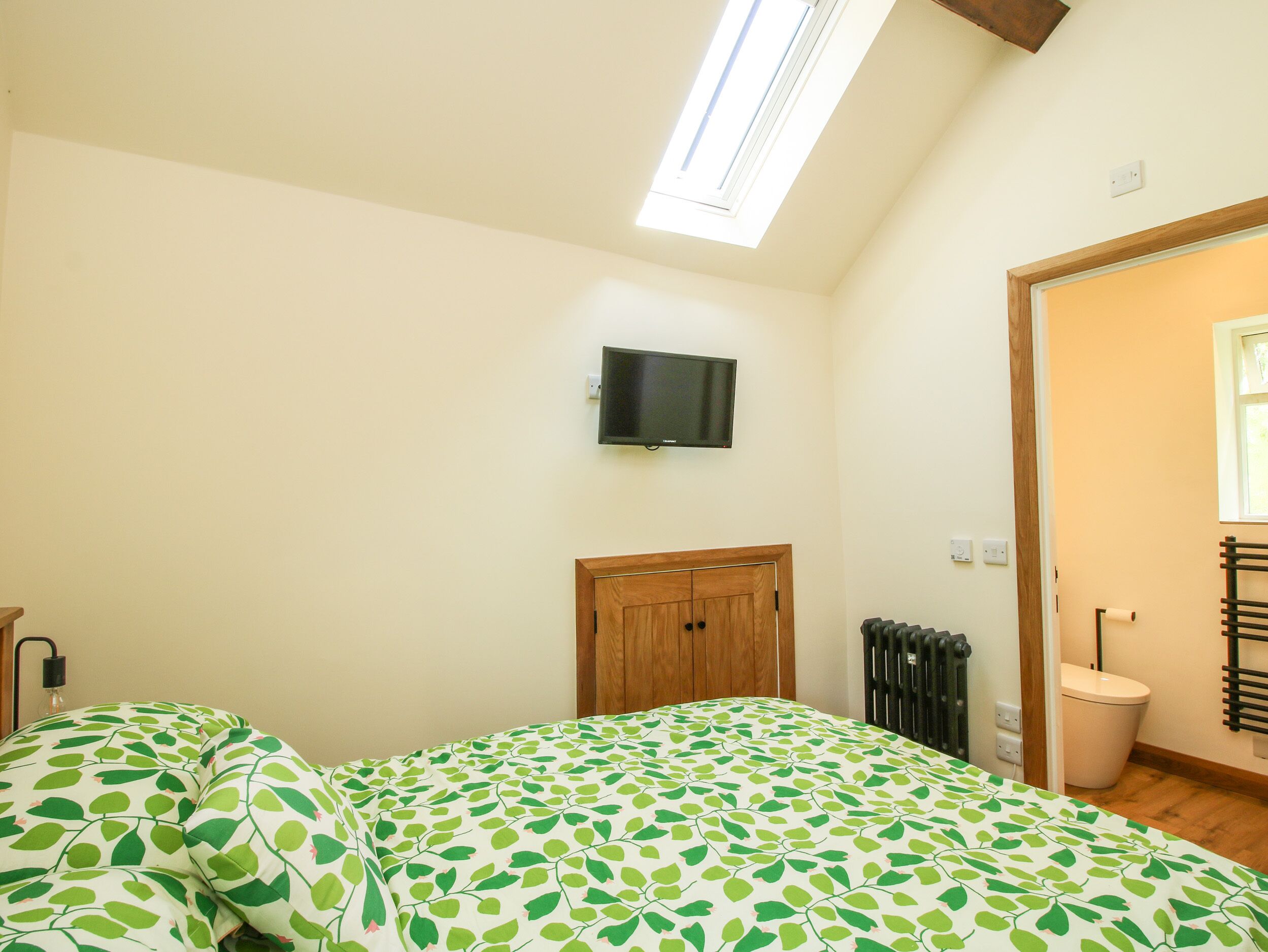 Quarto com cama, televisão na parede, radiador e porta que dá para outro espaço.
