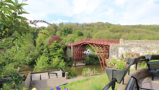 A red metal bridge over a river, surrounded by greenery and a stone wall. - The Office (Telford)