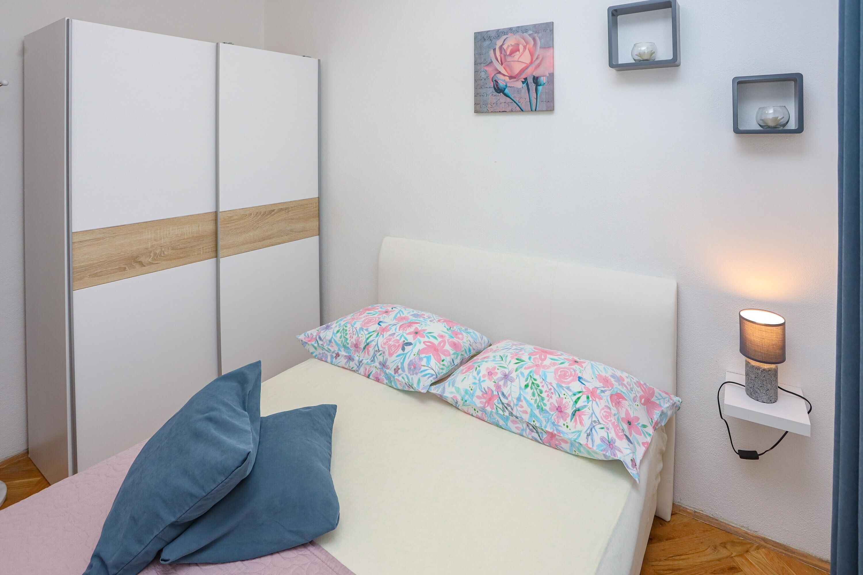 A bedroom with a bed, pillows, a wardrobe, a bedside table with a lamp, and a wall-mounted shelf with decorative items.