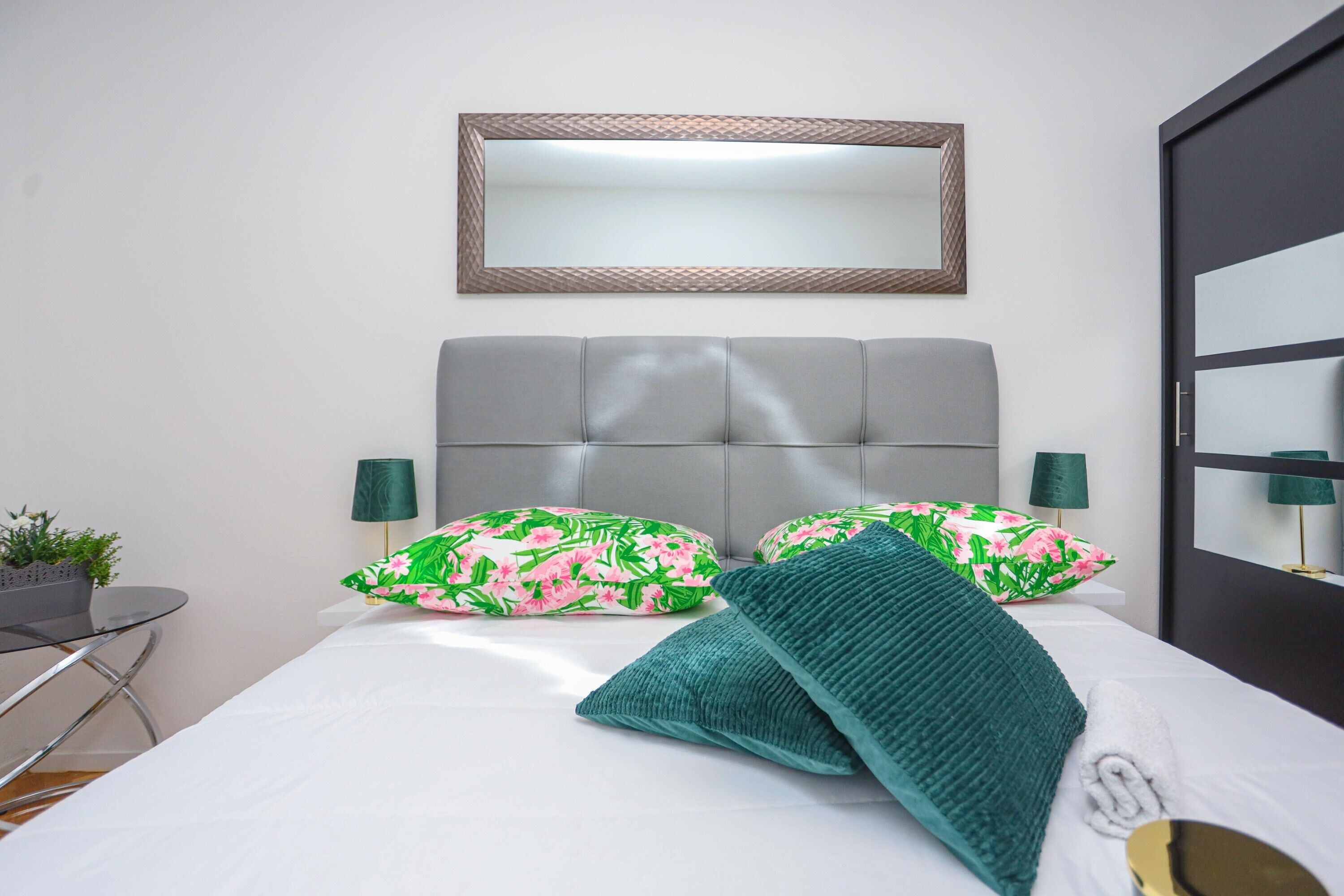 A neatly arranged bedroom with a bed, pillows, a bedside table, a lamp, and a mirror on the wall.