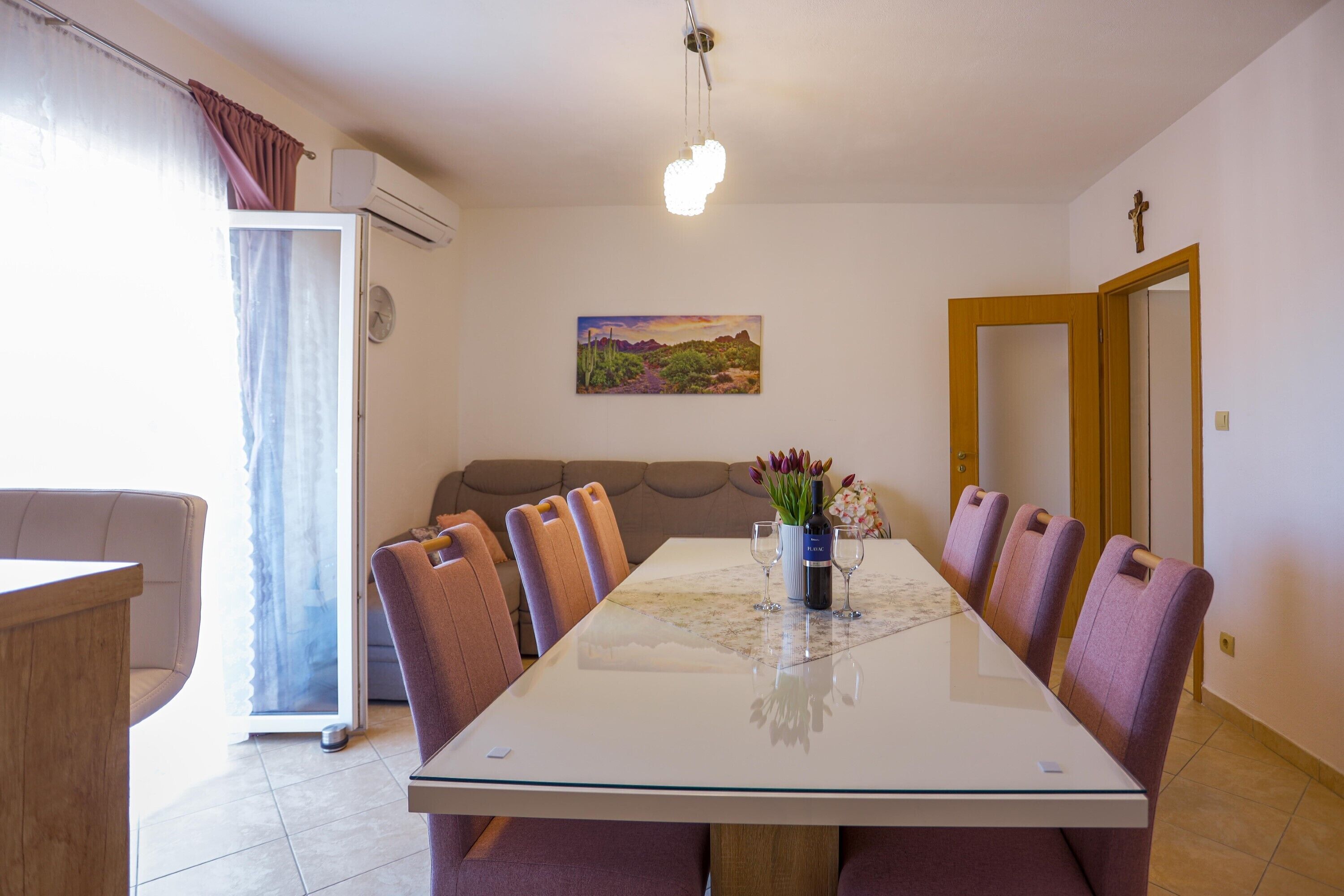 A dining room with a long table, six chairs, a vase with flowers, and a painting on the wall.