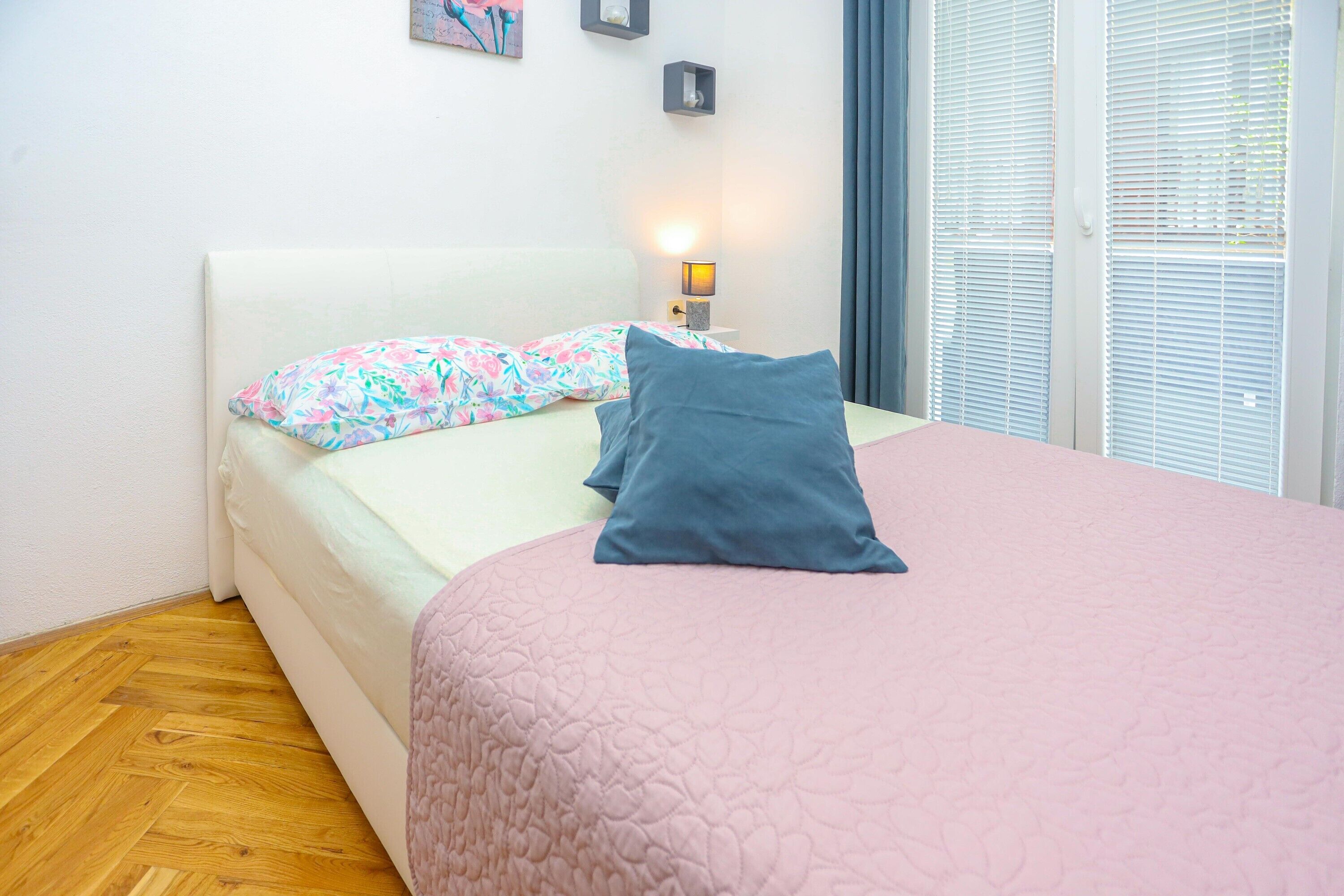 Una cama bien tendida con un edredón rosa y dos almohadas, una mesita de noche con una lámpara y una ventana con persianas.