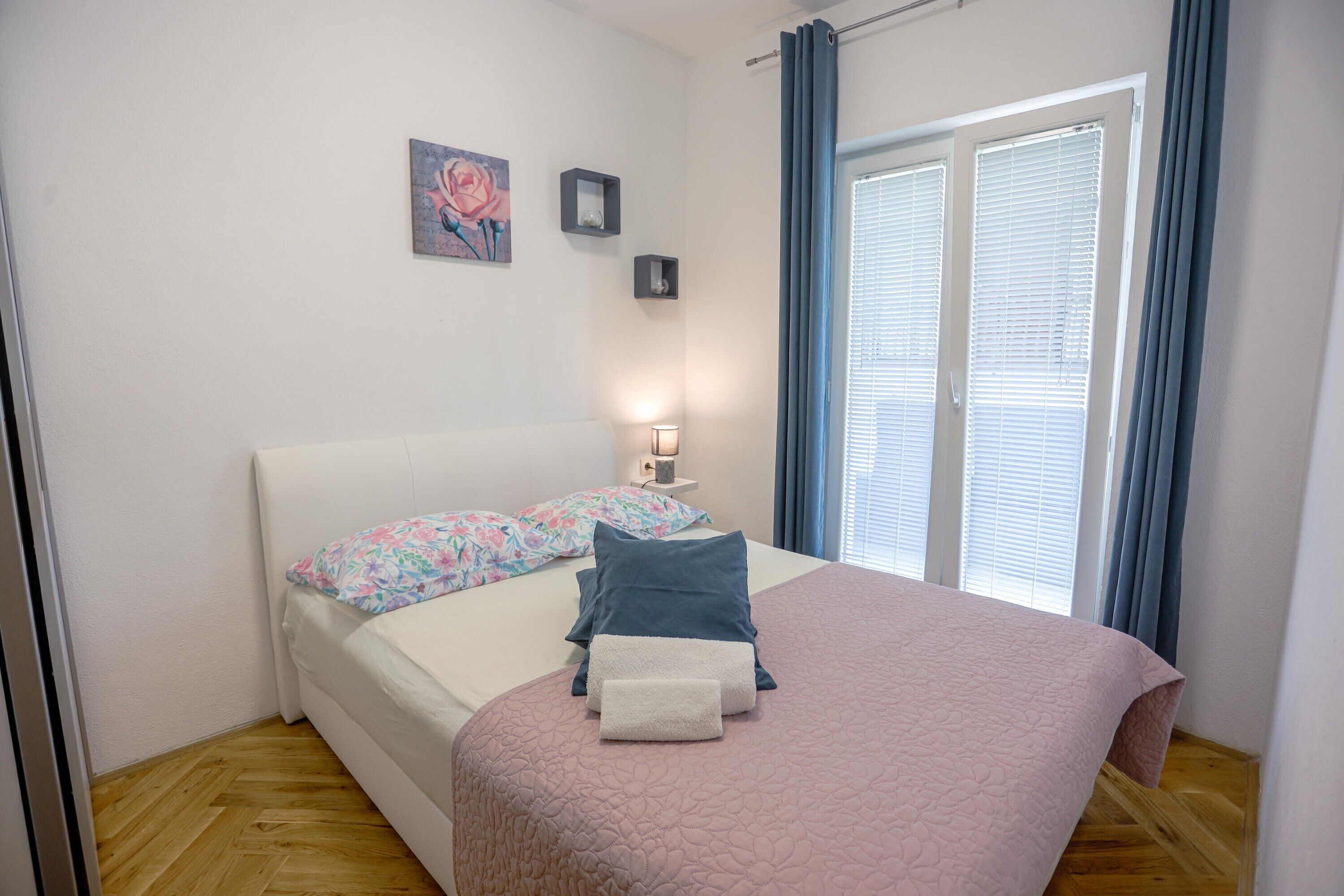 A bedroom with a bed, pillows, a bedside table with a lamp, a painting on the wall, and a window with curtains.