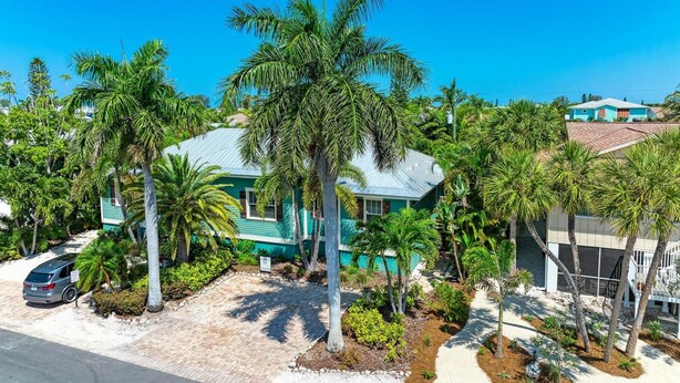A house with a blue roof, surrounded by palm trees and a paved driveway.