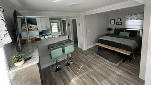 A modern bedroom with a bed, bedside table, and a small seating area. A kitchenette with a countertop and appliances is visible in the background. - Perfectly Small House (Xenia)