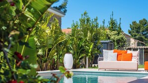 A pool area with a white sofa and orange cushions, surrounded by lush greenery and a clear blue sky.