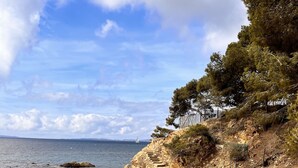 Una spiaggia rocciosa con acque cristalline, un sentiero sterrato e una scala che sale verso una scogliera.