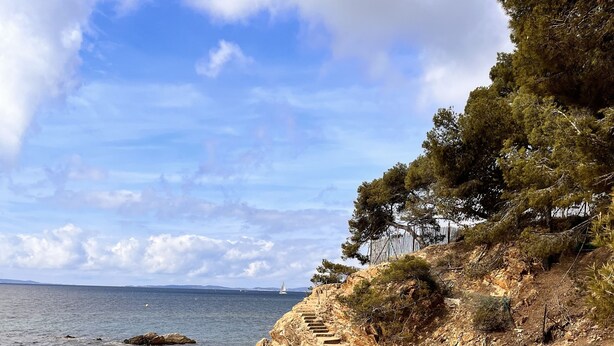 A rocky beach with clear water, a dirt path, and a staircase leading up a cliff.
