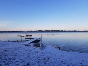 Lumipeitteinen ranta, josta ulottuu tyynen veden ylle puinen laituri. Rannalla on myös penkki ja kauempana näkyy puita reunustama rantaviiva.