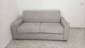 A plain, light-colored sofa in a corner of a room with tiled flooring and a plain white wall. - Newhouse Class (Bari)